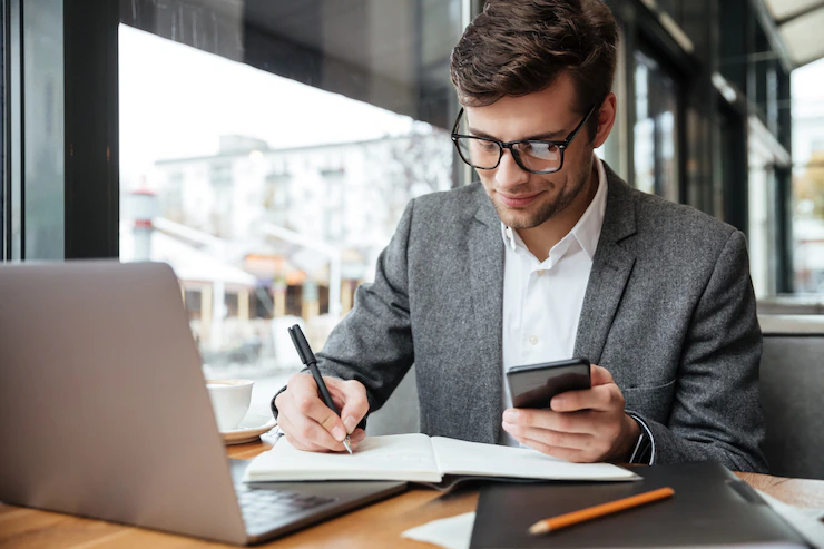 empresario sorridente em oculos sentado junto a mesa no cafe com o computador portatil enquanto estiver usando o smartphone e escrevendo algo 171337 5589
